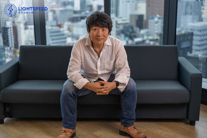 Developer Hideaki Itsuno sitting on a dark sofa in front of a large window