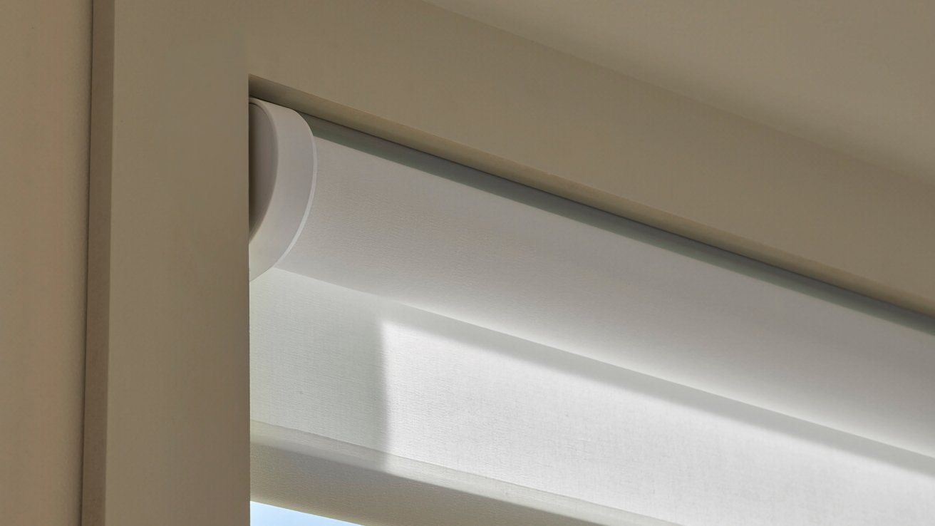 Close-up of a white roller blind partially extended, mounted within a beige window frame, with soft natural light casting gentle shadows.