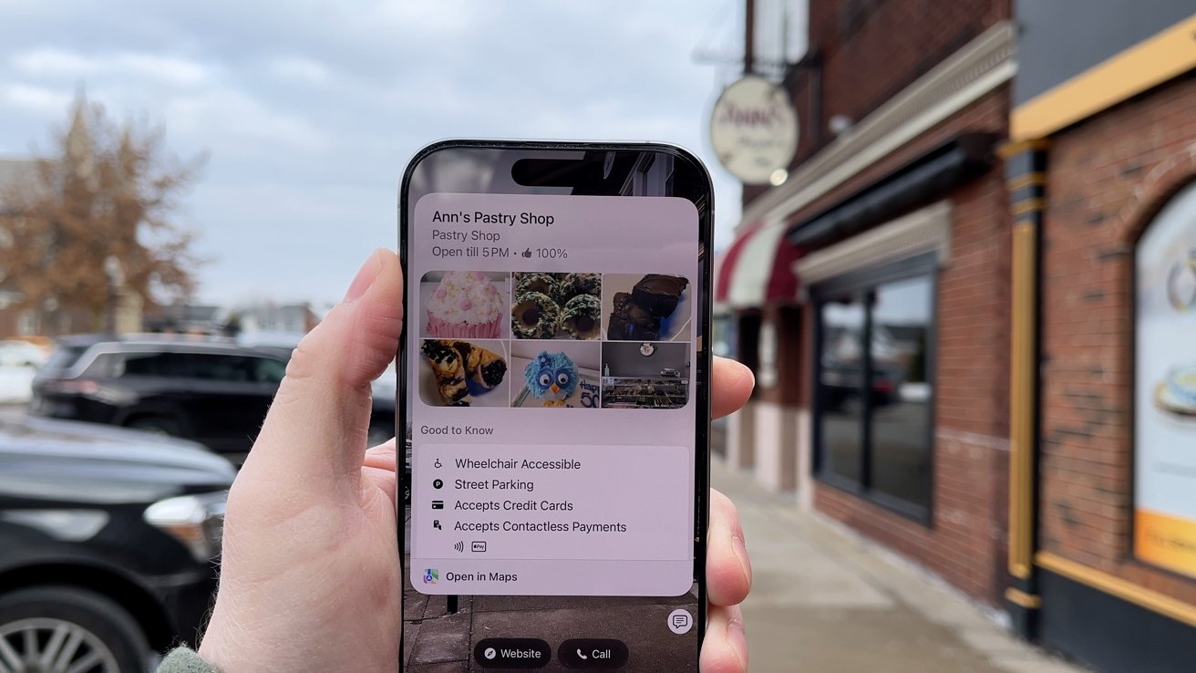 A hand holds a smartphone displaying bakery information, including photos and accessibility details, in front of a pastry shop on a cloudy street.