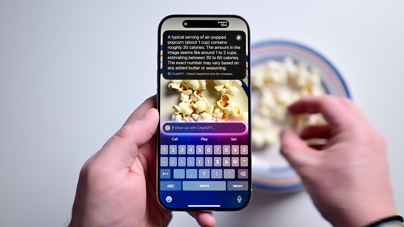 Person holding smartphone near a bowl of popcorn, screen displaying information and keyboard interface, one hand reaching for popcorn in the background.