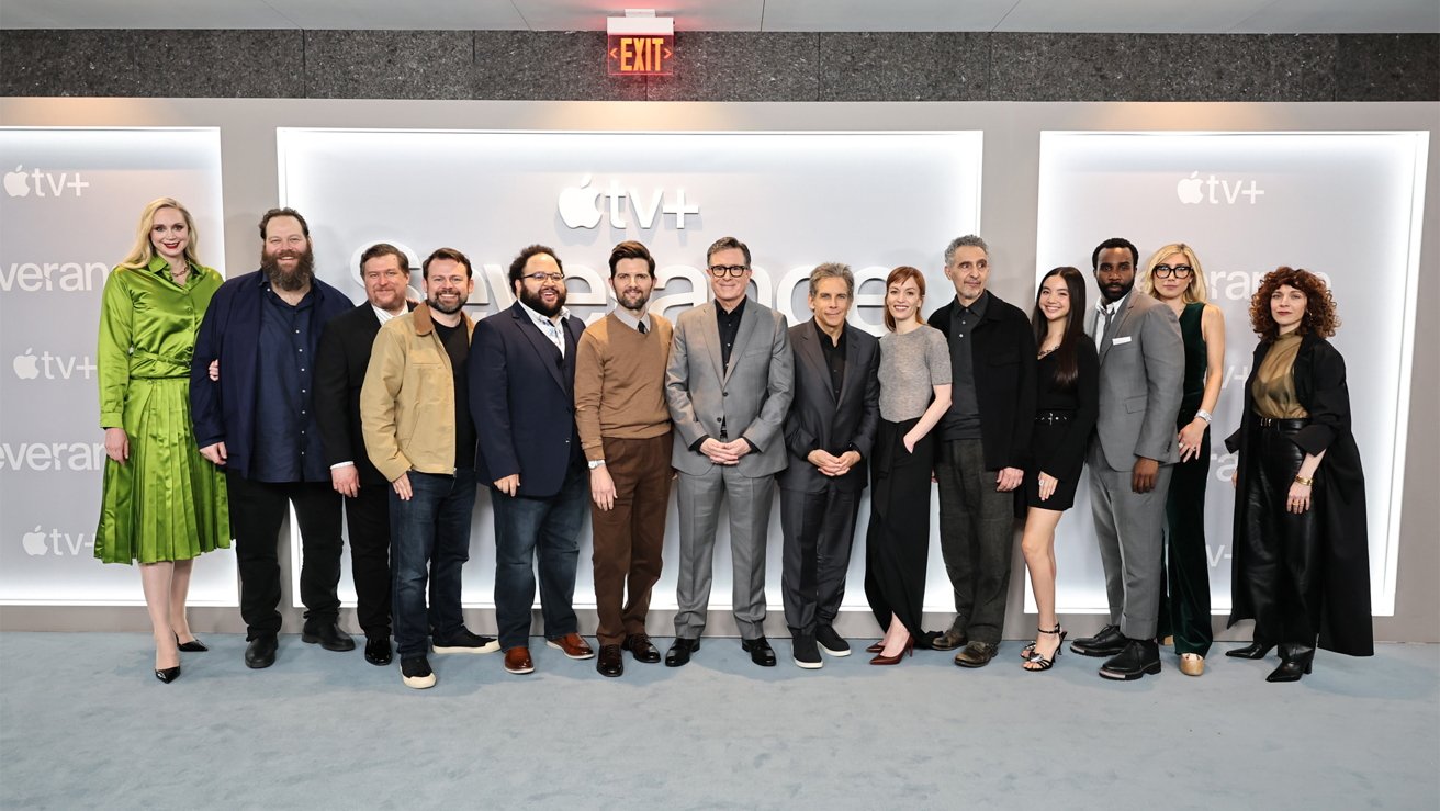 A group of 14 people, including 8 men and 6 women, stand together smiling in front of an Apple TV+ backdrop.