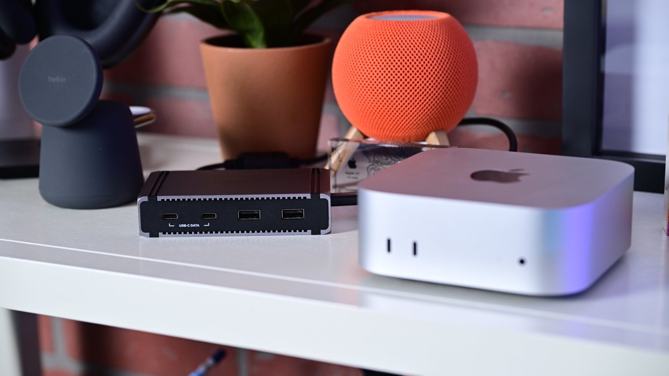 Desk with electronic devices, including a small silver computer, a black charging dock, an orange smart speaker, and a potted plant in the background.