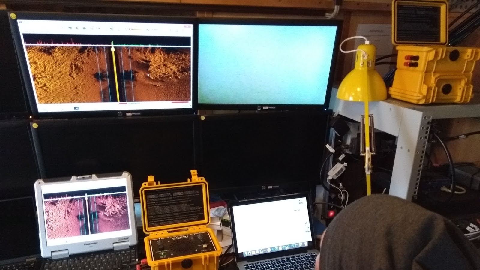 ghost-nets-fig-1 A photo of a workstation with four computer screens lit with sonar images. A man wearing a brown hat can be seen looking at the images, trying to identify ghost nets.